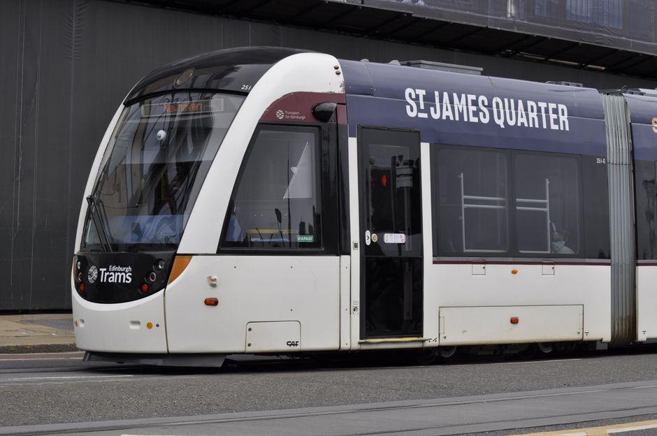 A modern tram operating in the St. James Quarter area, positioned on tracks next to a dark, contemporary building with a reflective surface. The tram has a sleek, white exterior with black-tinted wind